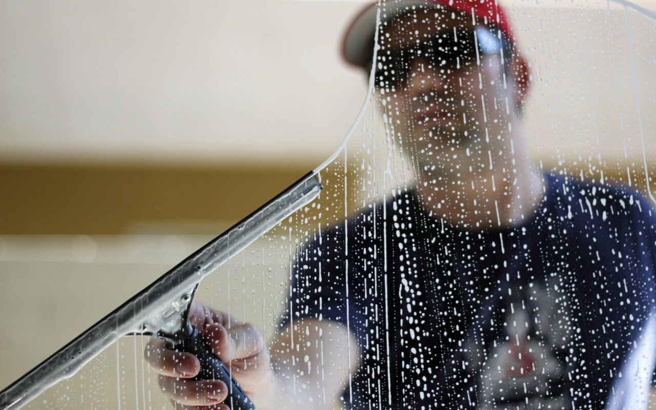 A close-up of a squeegee clearing soapy water from a residential window, water beading on glass.