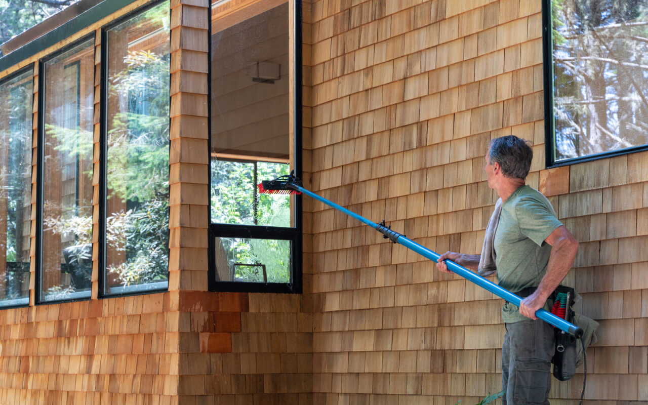 A crew member operating a tall water-fed pole on a residential cedar-shake exterior.